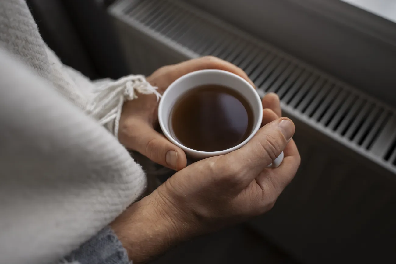 Person wrapped in blanket holding hot drink in front of radiator