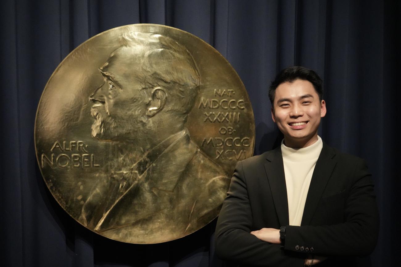 Max Han smiling with arms crossed, standing beside an enlarged replica of the Nobel Prize medal