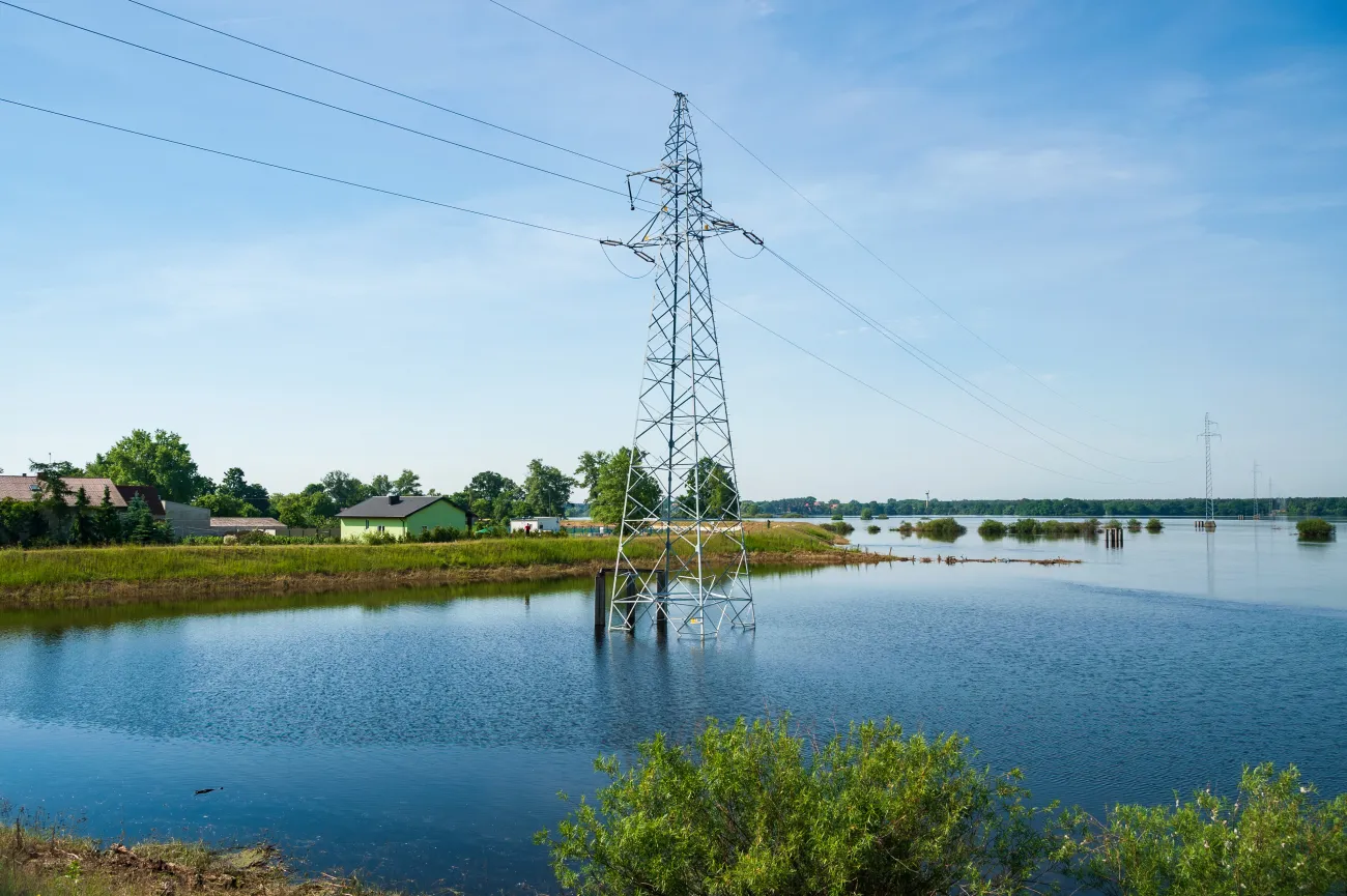 The high voltage power lines on water. Blue sky and green trees during river flood around a pylon tower