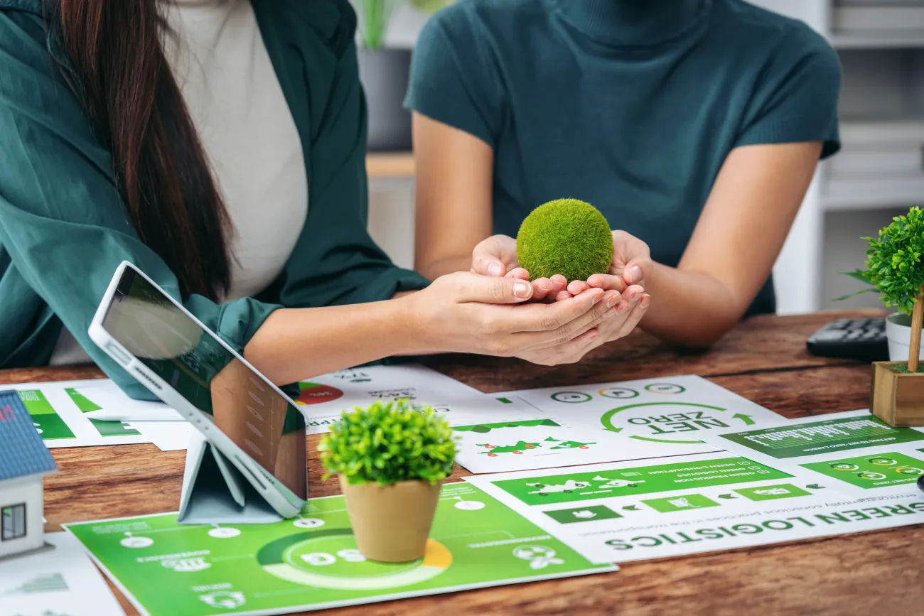 Two people discussing strategy while holding a green plant and looking at environmental images next to a laptop 