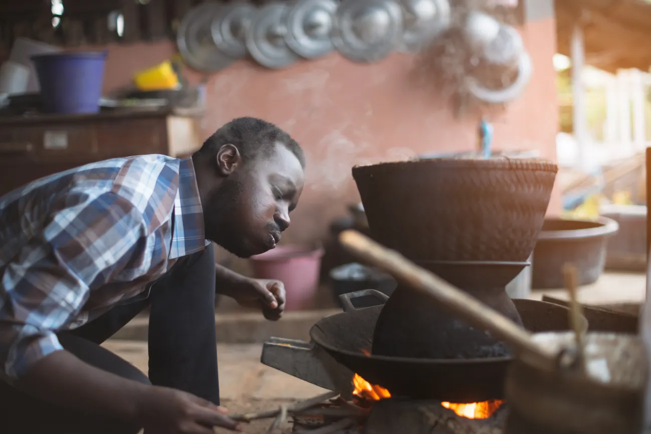 African man sitting down indoors and blowing a fire he's cooking rice on