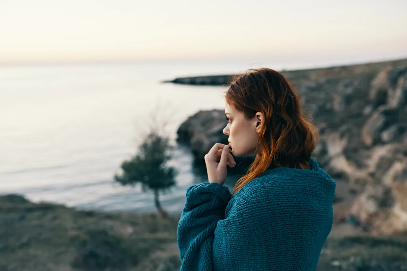 Young woman in mountains with blue scarf around shoulders, looking pensively at the sunset across the ocean