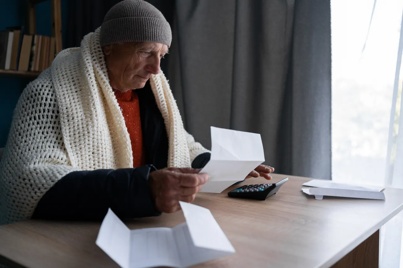 Senior man wrapped in blanket and wearing woolly hat opening UK energy bill concerned about cost of living energy crisis