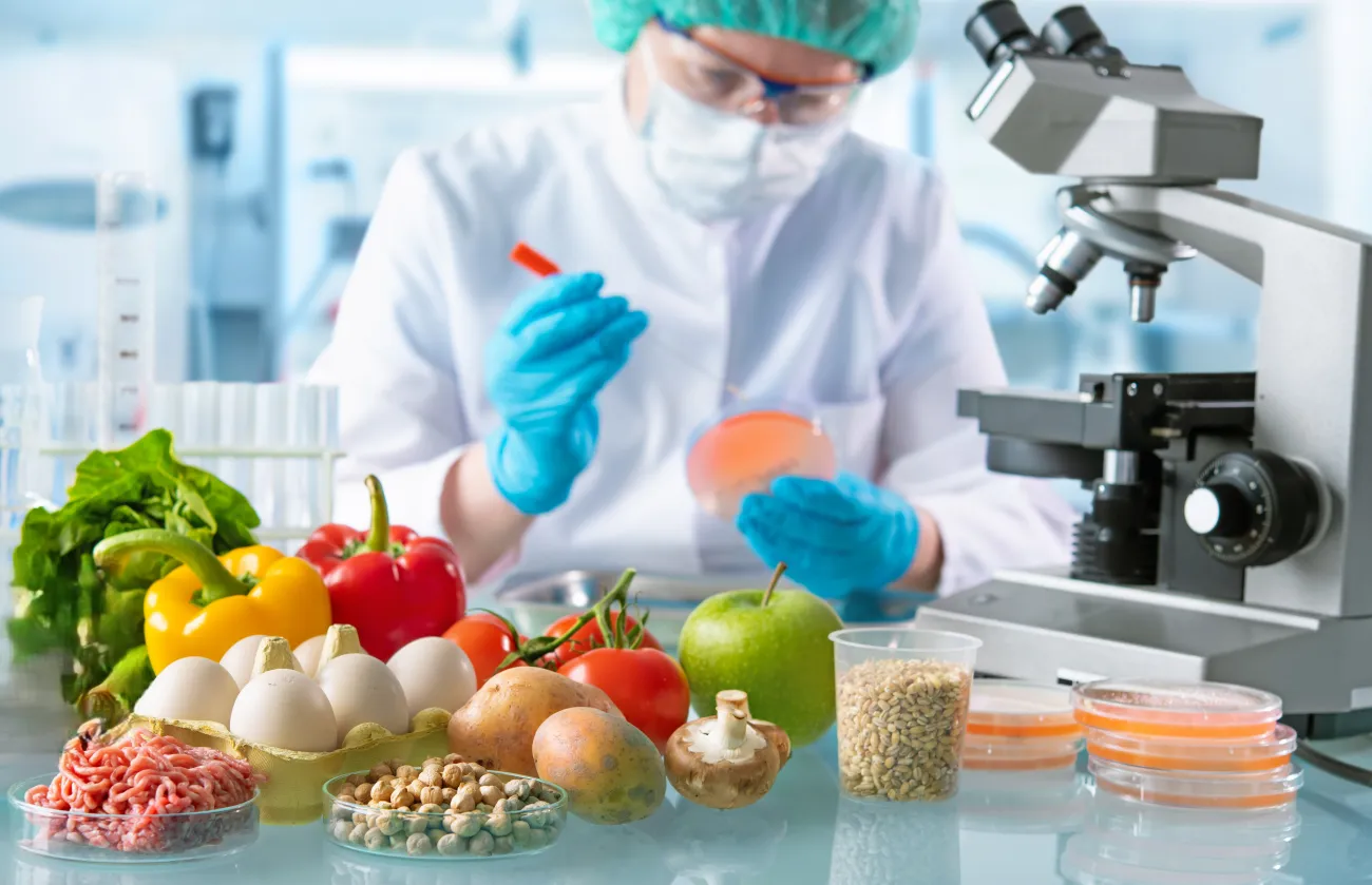 Scientist in lab coat, gloves and hair net studying food items in a lab