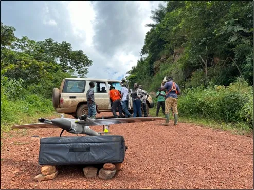 A drone in the foreground on a track near a forest with researchers getting equipment ready in the background