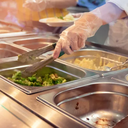 The gloved hand of a chef standing behind full lunch service station with assortment of food in trays holding some tongs